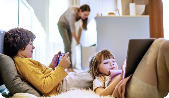 Bambini distesi che usano il tablet mentre la mamma sullo sfondo riordina la cucina.